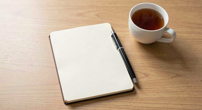 A blank notebook page with a pen and a cup of tea on a plain wooden table in soft daylight.