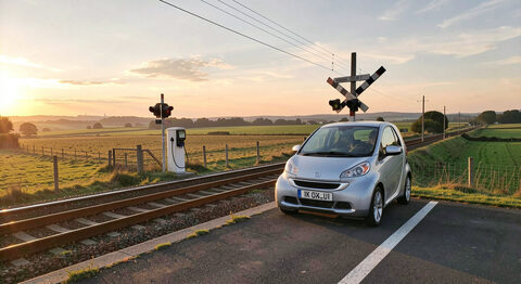 A small efficient car parked at a charging point beside a train line crossing open countryside at dawn